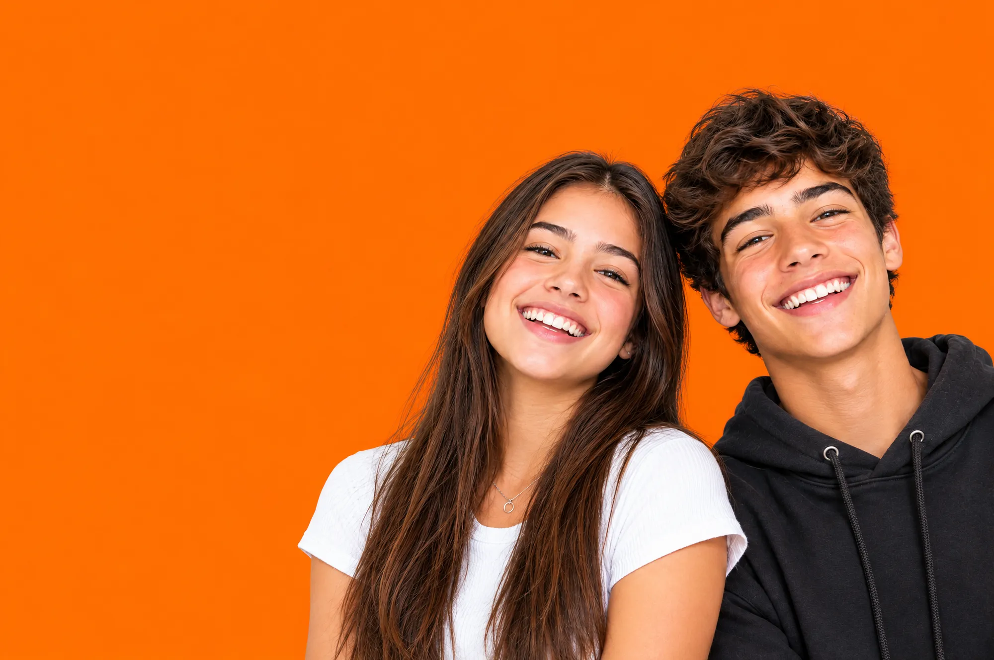 Dois adolescentes sorrindo lado a lado sobre fundo laranja, uma garota de cabelo longo e camiseta branca e um garoto de cabelo cacheado e moletom preto